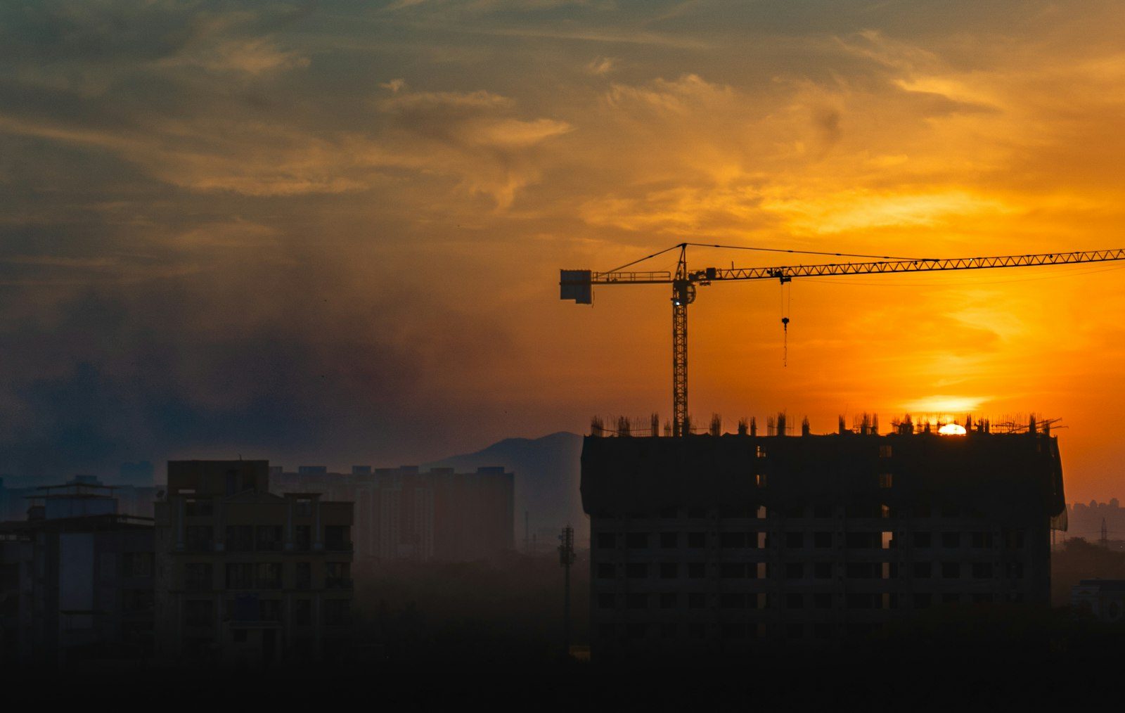Grua de torre sobre edifício com esperas de pilares no último pavimento, em silhueta ao pôr do sol